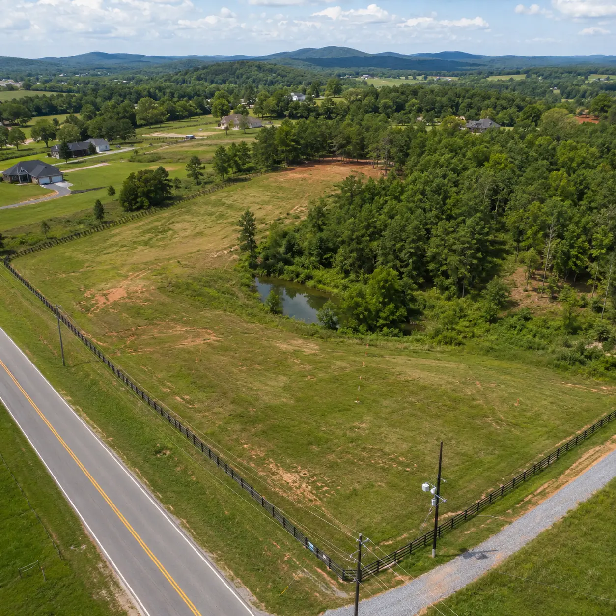 Aerial view of a rural land parcel showing road access, utility lines, nearby homes, trees, terrain, and a pond—key factors that can affect land value.
