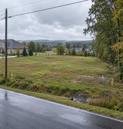 Vacant suburban land with road access, utility lines, drainage, survey markers, nearby development, and terrain features used by AI land valuation algorithms to estimate property value.